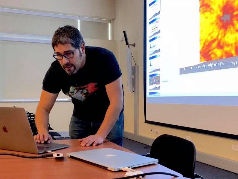 Bearded man in a black t-shirt leaning over a laptop, large screen in background displaying data.