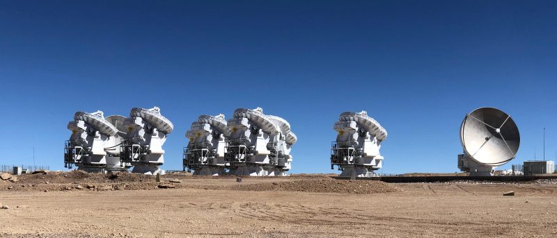 White radio telescopes with dishes on top above complicated machinery, under cobalt sky.