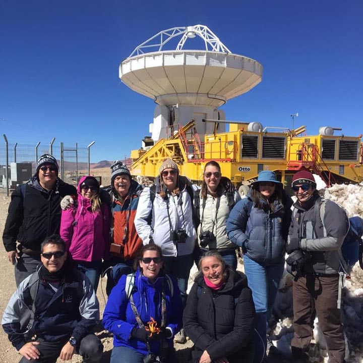 Ten people in coats with cameras posing in front of large radio telescope pointed straight up.