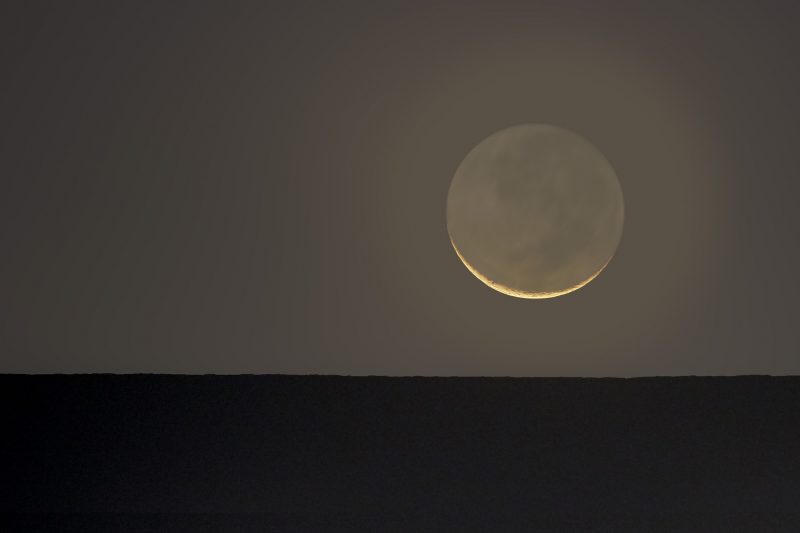 Thin, bright crescent with rest of moon dimly lit by earthshine above flat bare ground.