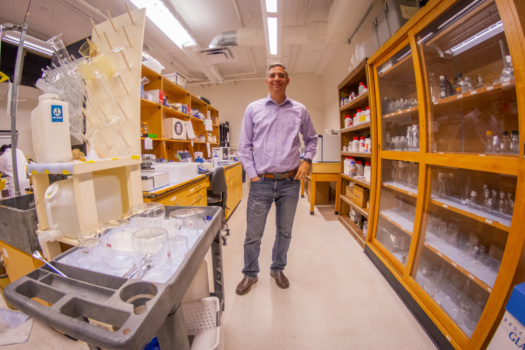 A man stands in an aisle between shelves and racks holding scientific equipment