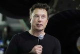 Elon Musk is wearing a black tee-shirt and speaking into a microwave at a public event