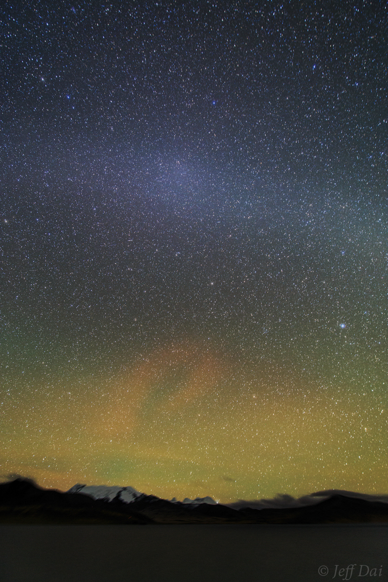 Night sky over mountainous horizon with fuzzy, pale white streak across it brighter in the middle.