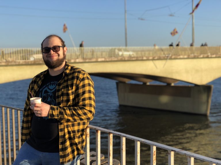 Smiling man with beard and sunglasses, with bridge in background.