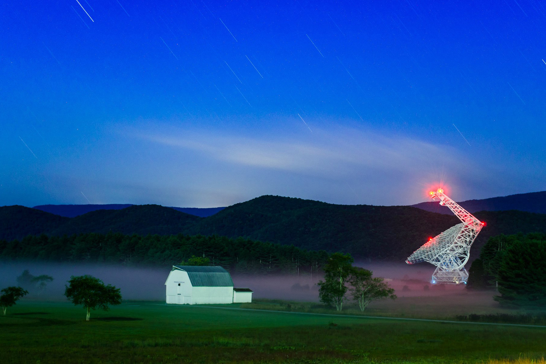 Photo with the Green Bank Telescope at right and a barnhouse at left, set in front of green mountains and a blue sky